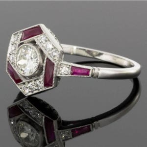 May include: A silver ring with a geometric design, featuring a central round diamond surrounded by smaller diamonds and rectangular rubies. The ring's band is smooth and polished, reflecting light.