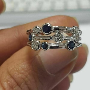 Può includere: Un set di tre anelli d'argento, ciascuno con una fila di pietre preziose rotonde alternate. Le pietre preziose includono diamanti trasparenti e zaffiri blu scuro. Gli anelli sono impilati, creando un effetto a strati.