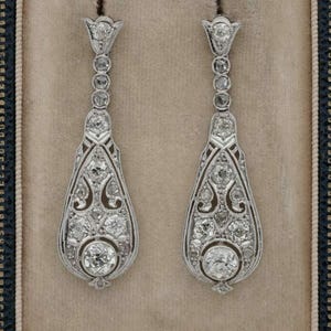 Può includere: Un paio di orecchini pendenti eleganti. Ogni orecchino presenta una serie di pietre preziose rotonde e scintillanti incastonate in un metallo color argento. Il design include una parte superiore floreale, una catena di pietre più piccole e una sezione a goccia.