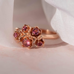 May include: A rose gold ring featuring a cluster of small, pink gemstone flowers. The ring's band is thin, and the floral design is the focal point. The gemstones are a vibrant pink color, and the ring is set against a soft, blurred background.
