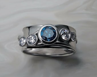 May include: A silver ring with a wide band, featuring a large, round, blue gemstone in the center, flanked by three smaller, clear gemstones. The ring has a modern, elegant design.