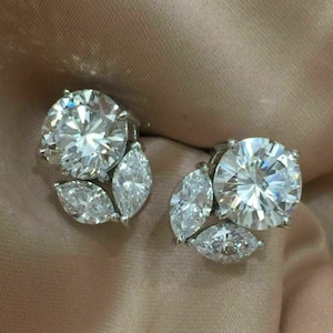 May include: A pair of sparkling stud earrings. Each earring features a large, round, clear gemstone surrounded by three smaller, marquise-cut stones. The earrings are set against a soft, peach-colored background.