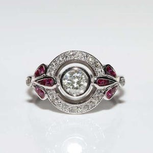 May include: A silver ring with a round, clear center stone surrounded by smaller diamonds. The ring has two wing-shaped accents on either side of the center stone, each accented with red rubies.