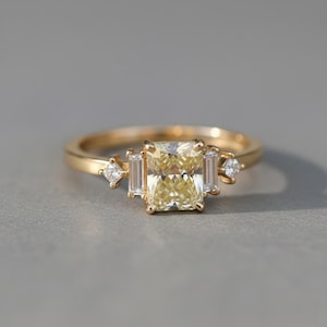 May include: A gold ring featuring a large, rectangular, yellow gemstone as the centerpiece, flanked by smaller, rectangular and round, clear gemstones. The ring has a delicate band and is set against a neutral background.