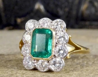 May include: A gold ring featuring a rectangular emerald center stone surrounded by a halo of round, brilliant-cut diamonds. The ring's setting has a vintage style, with a detailed band and a cluster of diamonds around the emerald.