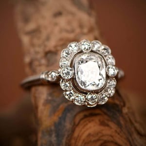 May include: A silver ring with a large, square-cut, clear gemstone surrounded by smaller, round gemstones. The ring is set on a wooden surface, showcasing its intricate design and sparkle.
