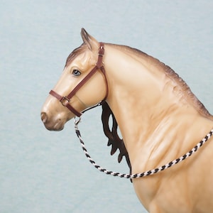 May include: A tan horse figurine wearing a brown leather halter and a black and white rope. The horse has a detailed mane and a light blue background. The horse is facing left.