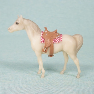 Puede incluir: Un caballo de juguete color crema con una silla de montar marrón. La silla tiene un asiento con estampado rosa y blanco. El caballo está sobre una superficie azul claro. El caballo de juguete es una réplica en miniatura de un caballo real.