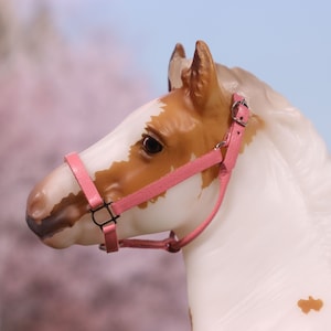 Puede incluir: Primer plano de un caballo de juguete blanco y marrón con un cabestro rosa. El caballo tiene una mancha marrón en el costado. El fondo está desenfocado y es rosa.