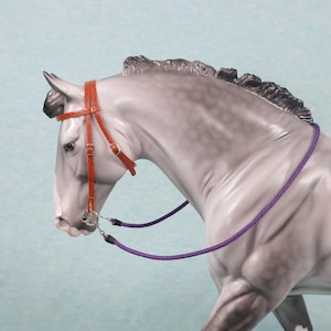 May include: A gray horse figurine with a red leather bridle and a purple rope. The horse is posed in profile, showcasing the detailed craftsmanship of the model. The bridle features buckles and straps, adding to the realism of the equestrian theme.