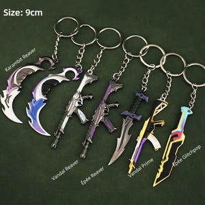 Può includere: Una collezione di portachiavi in metallo con vari disegni di armi, tra cui lame ricurve e fucili. I portachiavi misurano circa 9 cm e presentano una varietà di colori, tra cui argento, viola e oro. Il testo sull'immagine include "Karambit Reaver", "Vandal Reaver", "Épée Reaver", "Vandal Prime" e "Épée Glitchpop".