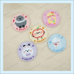 May include: A collection of five embroidered circular patches. Each patch features a different cartoon character, including a bird, a chick, an apple, a bunny, and a bear. Each patch has a name embroidered on it: Sylus, Rafael, Caleb, Xavier, and Zayne.