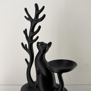 May include: A matte black cat figurine with bright green eyes, positioned next to a tall, branching tree-like structure. The cat's tail extends to support a small, circular, bowl-shaped tray, creating a functional display piece.