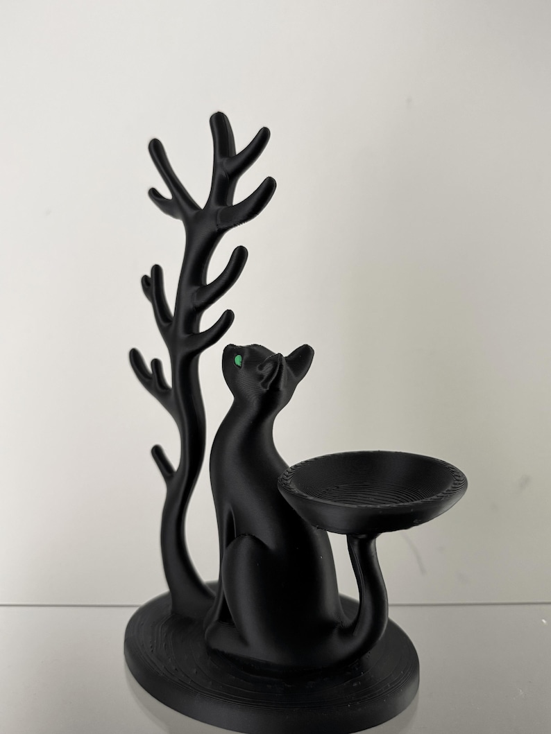 May include: A matte black cat figurine with bright green eyes, positioned next to a tall, branching tree-like structure. The cat's tail extends to support a small, circular, bowl-shaped tray, creating a functional display piece.