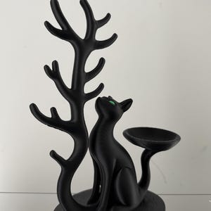 May include: A matte black cat figurine with bright green eyes, sitting on a circular base. To the left, a tall, branching tree-like structure rises from the base. To the right, a small, shallow, circular dish is attached to the cat's tail.
