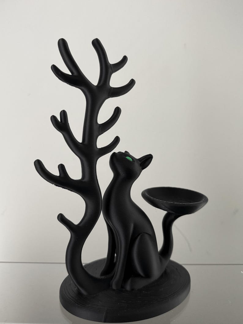 May include: A matte black cat figurine with bright green eyes, sitting on a circular base. To the left, a tall, branching tree-like structure rises from the base. To the right, a small, shallow, circular dish is attached to the cat's tail.