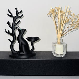 May include: A matte black cat figurine with a green eye, featuring an attached tree-like branch and a small circular dish. Beside it sits a clear glass reed diffuser bottle containing light-coloured reeds and dried beige flowers.