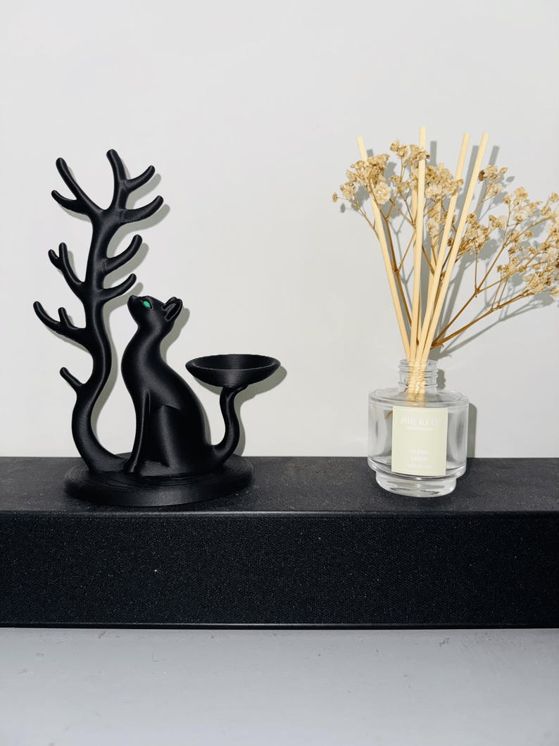 May include: A matte black cat figurine with a green eye, featuring an attached tree-like branch and a small circular dish. Beside it sits a clear glass reed diffuser bottle containing light-coloured reeds and dried beige flowers.