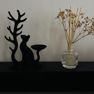 May include: A matte black cat-shaped candle holder with a tree-like branch design next to a clear glass reed diffuser bottle containing light brown wooden reeds and dried white flowers with a label reading AIR-KEEP CLEAN LINEN.