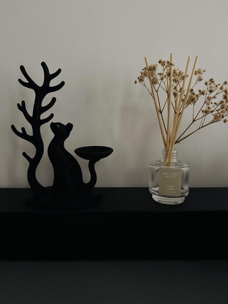 May include: A matte black cat-shaped candle holder with a tree-like branch design next to a clear glass reed diffuser bottle containing light brown wooden reeds and dried white flowers with a label reading AIR-KEEP CLEAN LINEN.