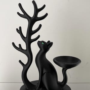 May include: A matte black cat figurine with bright green eyes, positioned next to a tall, branching tree-like structure on one side and a small, circular, bowl-shaped tray extending from the cat's tail on the other side.