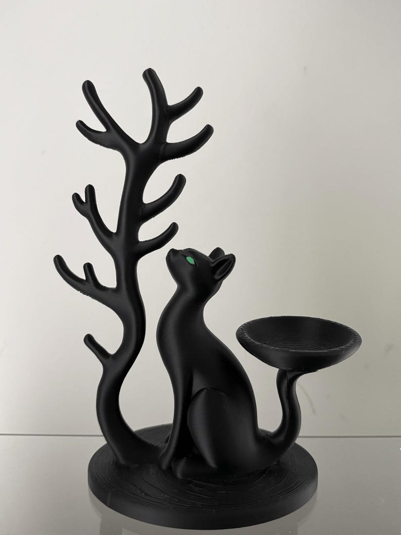 May include: A matte black cat figurine with bright green eyes, positioned next to a tall, branching tree-like structure on one side and a small, circular, bowl-shaped tray extending from the cat's tail on the other side.