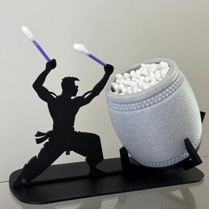 May include: A novelty cotton bud holder featuring a black silhouette of a person playing a drum. The drum is grey and filled with cotton buds. The person is holding two purple-tipped cotton buds as drumsticks.