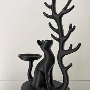 May include: A matte black jewellery stand featuring a seated cat with bright green eyes. The cat is positioned next to a tall, branching tree-like structure designed for hanging items, and a small circular dish extends from the cat's tail.
