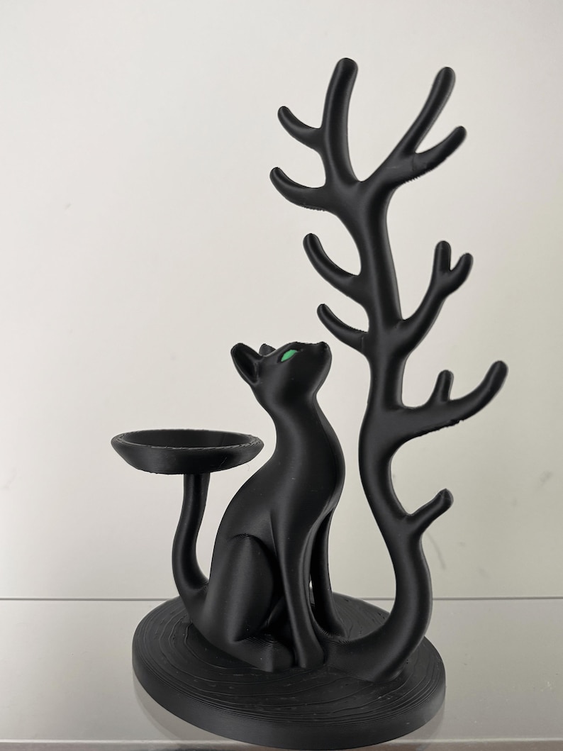 May include: A matte black jewellery stand featuring a seated cat with bright green eyes. The cat is positioned next to a tall, branching tree-like structure designed for hanging items, and a small circular dish extends from the cat's tail.
