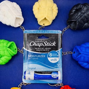 以下が含まれることがあります： SPF15のチャップスティックリップクリームのパッケージ。カラフルなミニチュアジャケットのキーホルダーに囲まれています。リップクリームのパッケージには「MOISTURIZER 2 in 1 Lipcare」と「SPF 15」の文字。キーホルダーは白、黄色、黒、青、赤、緑です。