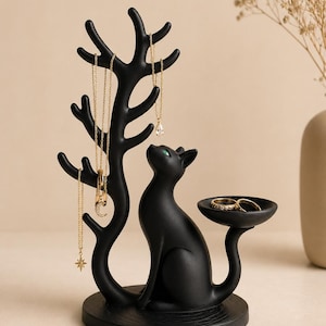 May include: A matte black jewellery stand featuring a cat figure sitting on a circular base. The cat has green eyes and a tail that extends into a small dish. A tree-like structure with multiple branches rises from the base to hold necklaces.