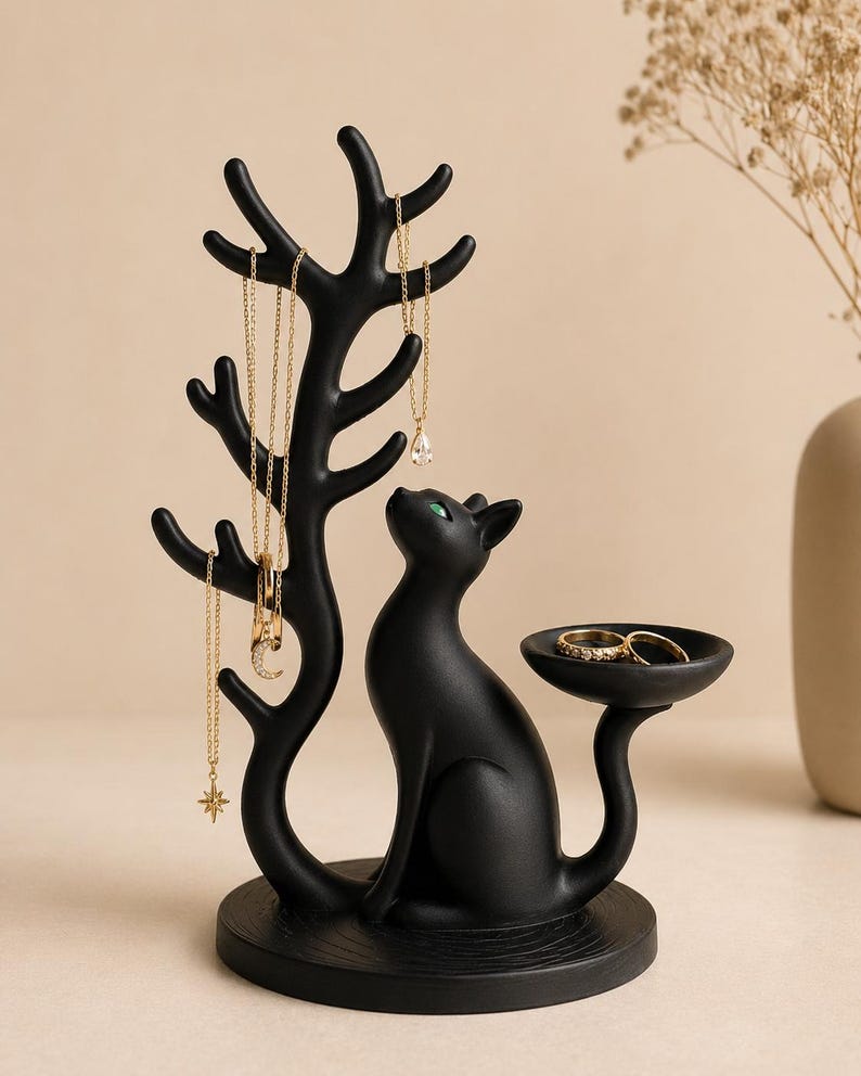 May include: A matte black jewellery stand featuring a cat figure sitting on a circular base. The cat has green eyes and a tail that extends into a small dish. A tree-like structure with multiple branches rises from the base to hold necklaces.