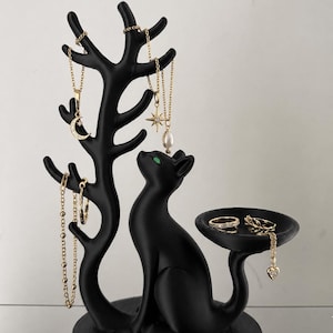 May include: A matte black jewellery organiser featuring a cat figure sitting beside a tree-shaped stand with multiple branches for hanging necklaces and bracelets, and a small circular tray for rings, accented with bright green cat eyes.