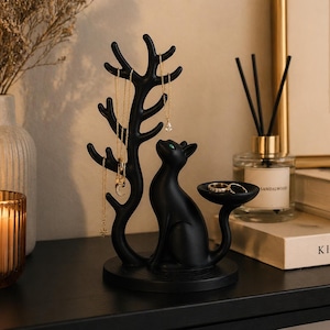 May include: A matte black jewellery organiser featuring a stylised cat sitting next to a tree-like structure with multiple branches for hanging necklaces. The cat has green eyes and a small circular tray attached to its tail for holding rings.