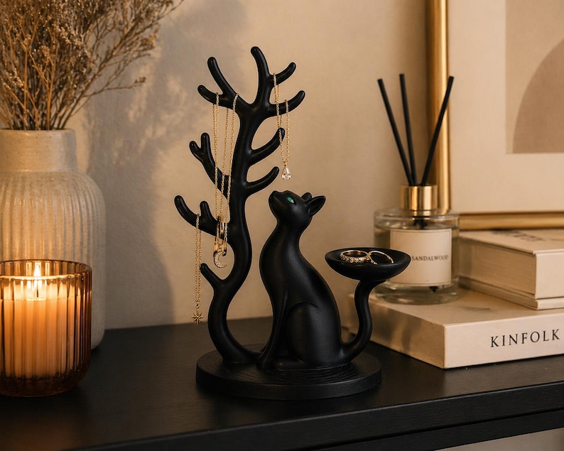 May include: A matte black jewellery organiser featuring a stylised cat sitting next to a tree-like structure with multiple branches for hanging necklaces. The cat has green eyes and a small circular tray attached to its tail for holding rings.