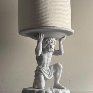 May include: A white toilet paper holder featuring a kneeling figure with a muscular build, wearing a draped cloth around the waist, holding a circular platform above the head to support a single roll of white toilet paper.