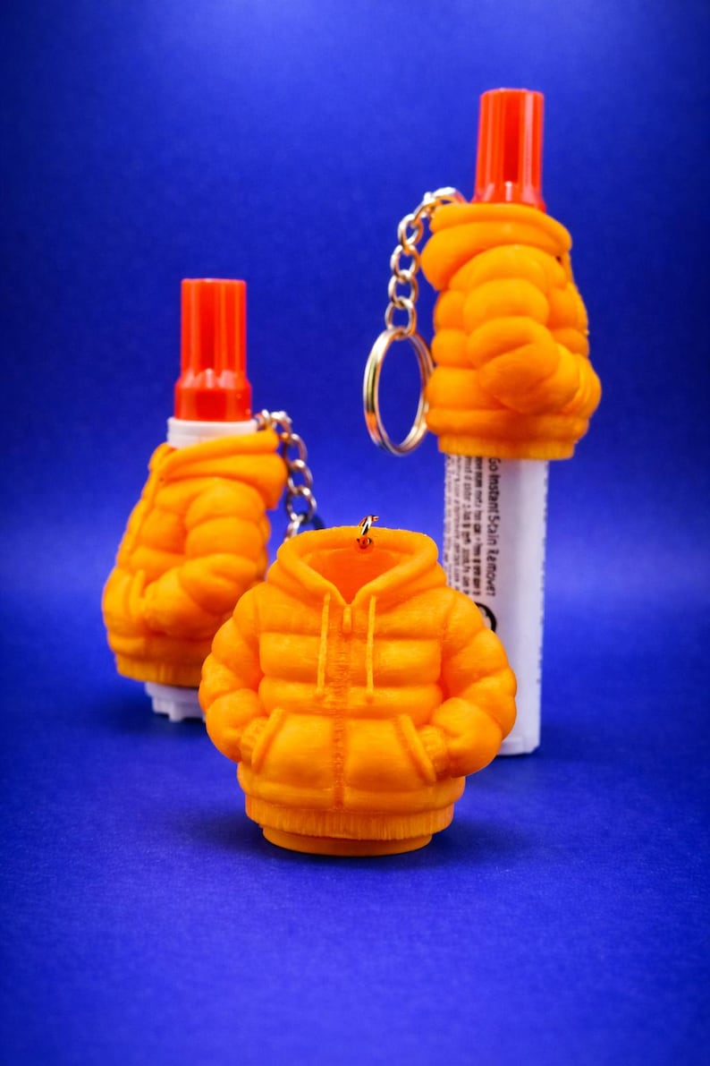 Orange puffer jacket charm holding a mini stain remover pen, on a keyring over warm wood