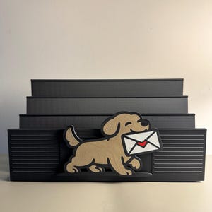 May include: A black tiered desk organiser featuring a tan dog figure holding a white envelope with a red heart in its mouth. The organiser has four horizontal slots for holding mail or documents, with a textured, ribbed design on the front panel.