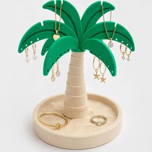 May include: A palm tree-shaped jewellery organiser with a beige circular base and a textured trunk. The green palm fronds feature small holes for hanging earrings and necklaces. A gold bangle and a small ring rest on the sandy-textured base.