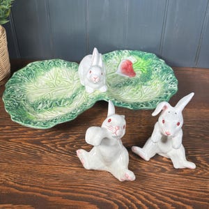 May include: A ceramic Easter serving dish and bunny figurines. The green dish is shaped like a cabbage leaf with a red accent. Three white bunny figurines with pink accents are arranged around the dish, creating a charming spring display.