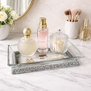 May include: A rectangular glass tray with a silver floral design displays perfume bottles, a small glass jar, and jewellery. The tray has a clear glass top and a silver base. A gold-rimmed mirror and a vase of white flowers are in the background.
