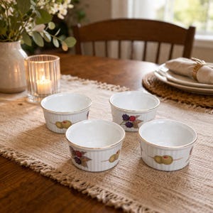 May include: Four white ceramic ramekins with gold rims and vertical ribbed sides. Each ramekin features a different fruit design: olives, blackberries, and raspberries. The ramekins are arranged on a beige table runner.
