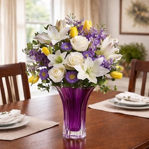 May include: A vibrant floral arrangement in a purple glass vase. The bouquet features white lilies, white roses, yellow tulips, and purple and yellow accent flowers. The vase is on a wooden table, with place settings visible.