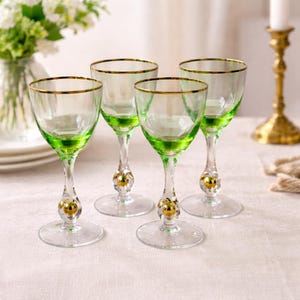 May include: Four clear wine glasses with green bases and gold rims. Each glass has a decorative gold ball on the stem. The glasses are arranged on a white tablecloth, with a vase of flowers in the background.
