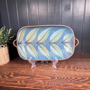 May include: Rectangular serving tray with a blue background and a leaf design in shades of green and yellow. The tray has brown handles and a brown border. The tray is displayed on a clear stand on a wooden surface.