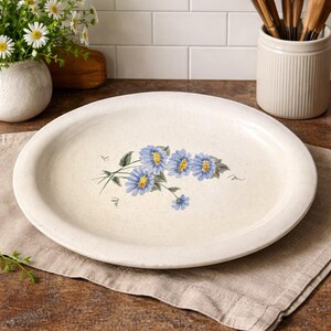 Könnte beinhalten: Cremefarbene ovale Servierplatte mit floralem Design. Die Platte zeigt einen Strauß blauer Blumen mit gelben Zentren und grünen Blättern. Die Blumen sind auf die cremefarbene Oberfläche gemalt. Der Rand der Platte ist leicht erhöht.