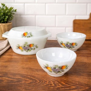 May include: A set of three white ceramic bowls, one with a lid, featuring a fruit design. The bowls have a glossy finish and are decorated with illustrations of oranges, pears, and grapes. The bowls are arranged on a wooden surface.