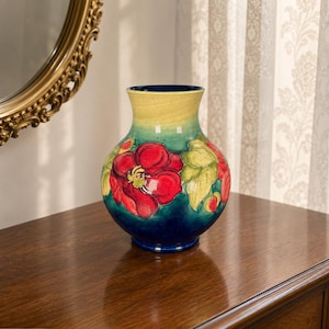 May include: A ceramic vase with a bulbous body and a flared rim, featuring a hand-painted design of large red flowers and green leaves against a gradient background that transitions from a deep blue base to a pale yellow top section.