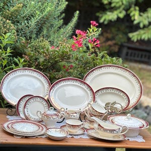 May include: A vintage porcelain dinner service in white with a burgundy and gold floral design. The set includes plates, serving dishes, teacups, a teapot, a gravy boat, and a lidded serving bowl. The items are arranged on a wooden table, with a lace doily.