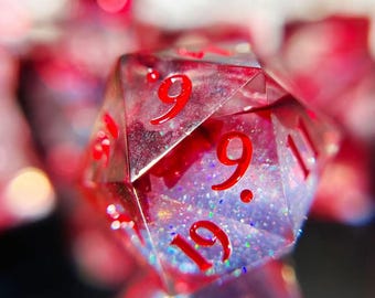 Puede incluir: Primer plano de un dado transparente de múltiples caras con números rojos. El dado tiene un interior rojo y morado con purpurina. Los números 9, 19 y 11 son visibles. El fondo está borroso con bokeh rojo y rosa.
